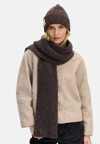 Brązowa dziergana czapka z prążkowaną teksturą, połączona z długim, pasującym brązowym szalem. Strój uzupełnia jasnobeżowa kurtka o fakturowanej powierzchni.