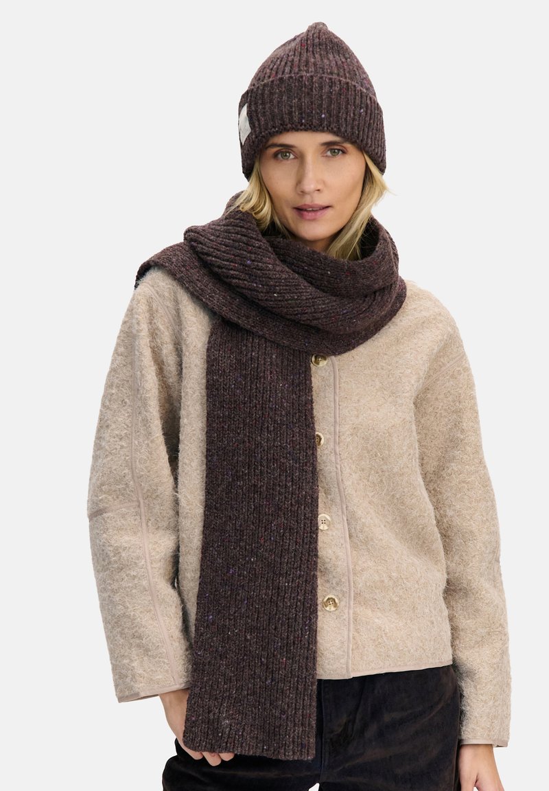 Brązowa dziergana czapka z prążkowaną teksturą, połączona z długim, pasującym brązowym szalem. Strój uzupełnia jasnobeżowa kurtka o fakturowanej powierzchni.