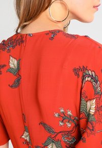 Kvinde iført store hoops øreringe og en rød blomstermønstret bluse med lynlås i ryggen, vist fra skulderen og op.