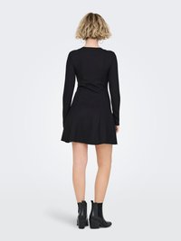 Robe noire à manches longues avec un corsage ajusté et une jupe évasée, associée à des bottines noires à talons. Tissu lisse, design simple.