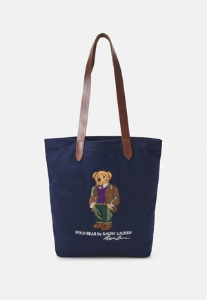 Sac cabas bleu marine avec des anses en cuir marron, présentant un ours brodé portant une veste, un pull et un pantalon avec le texte Ralph Lauren en dessous.