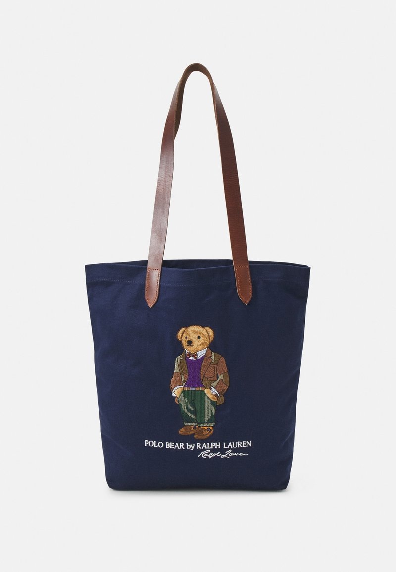 Sac cabas bleu marine avec des anses en cuir marron, présentant un ours brodé portant une veste, un pull et un pantalon avec le texte Ralph Lauren en dessous.