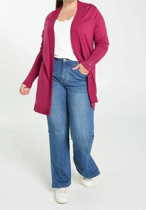 Femme portant un cardigan rose vif, un haut blanc, un jean bleu et des baskets blanches, debout avec une main dans sa poche.