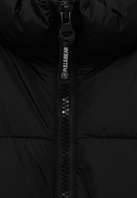 Veste noire matelassée avec une fermeture éclair à l'avant munie d'une tirette à logo et une texture matelassée, mettant en valeur son design isolant.