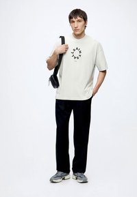 Νέος άνδρας φορώντας λευκό T-shirt με στάμπα, μαύρο παντελόνι, γκρι αθλητικά παπούτσια και μαύρη τσάντα χιαστί, στέκεται μπροστά σε απλό λευκό φόντο.