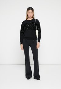 Sweatshirt preto com frente de lantejoulas, combinado com calças pretas à boca de sino. O modelo segura uma pequena bolsa branca, de pé contra um fundo simples.