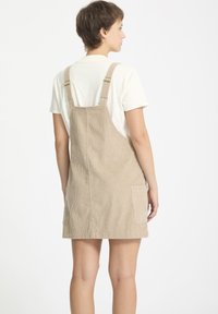 Beige corduroy tuinbroekjurk met verstelbare banden, voorzak en een gestructureerde A-lijn vorm. Draag over een wit T-shirt.