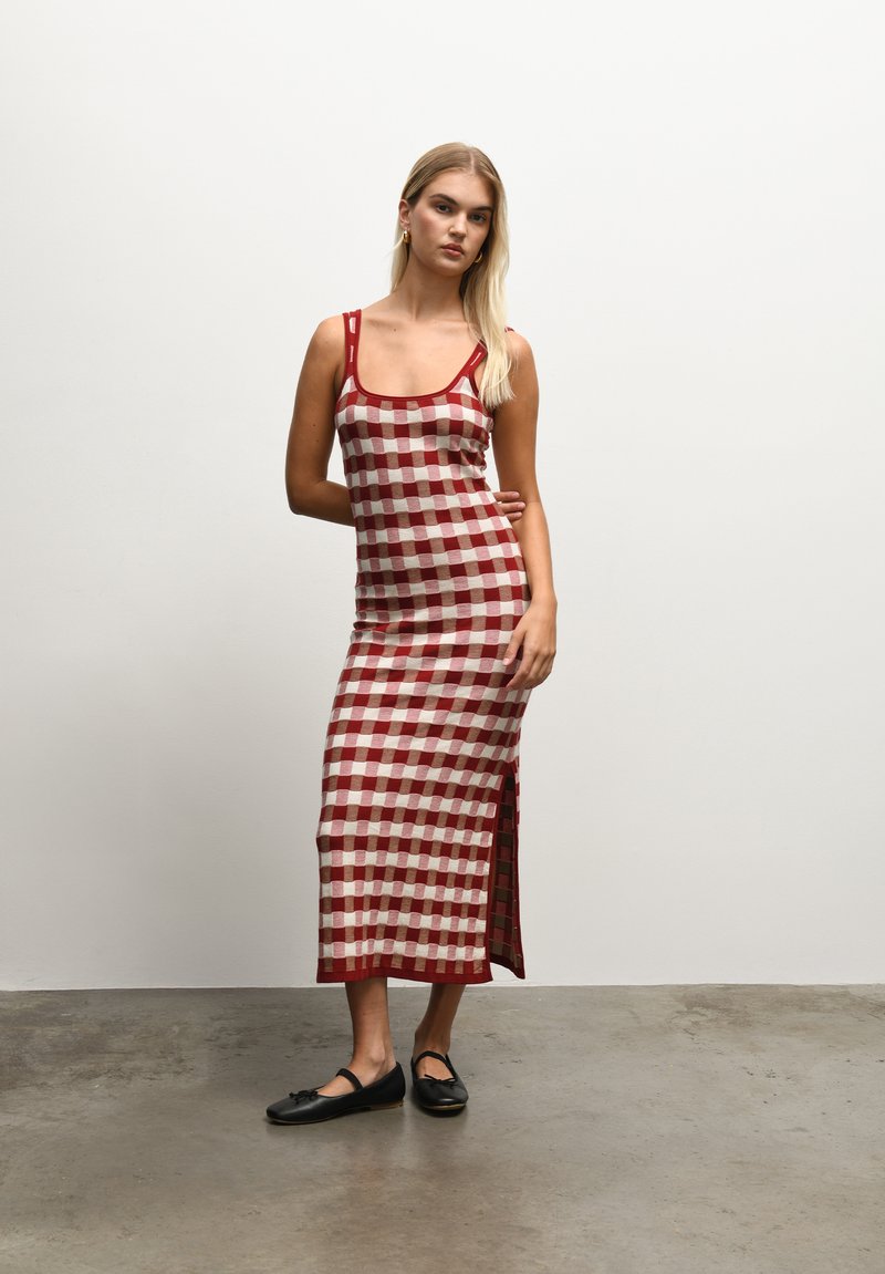 robe maxi à carreaux rouge et blanc, ajustée avec des bretelles fines, fente sur le côté, en tissu léger. Portée avec des chaussures plates noires.