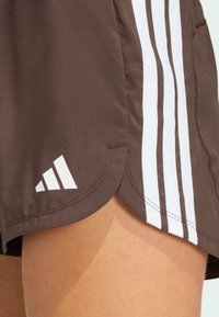 Shorts de sport marron avec design à trois bandes blanches, possédant un ourlet fendu et un logo Adidas blanc sur le côté inférieur gauche. Tissu texturé.