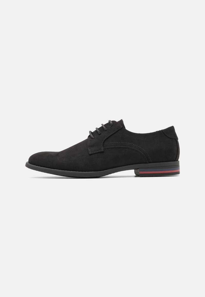 Chaussure habillée en daim noir pour homme avec lacets et talon bas présentant des rayures grises et rouges sur le bord de la semelle, vue de profil latéral sur fond blanc.