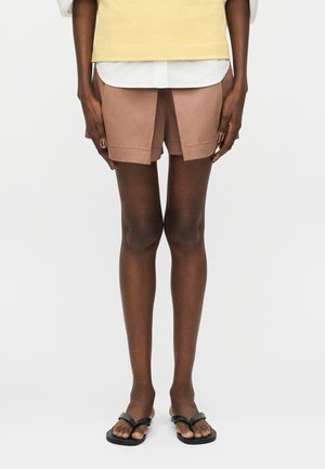 Άτομο που φοράει μπεζ σορτς, λευκό πουκάμισο, κίτρινο γιλέκο και μαύρα σανδάλια, στέκεται με τα χέρια χαλαρά δίπλα στο σώμα πάνω σε λευκό φόντο.