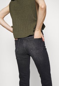 Jeans skinny gris foncé avec une poche arrière, associés à un haut sans manches côtelé vert olive. Le haut a une finition texturée et une fente sur le côté.