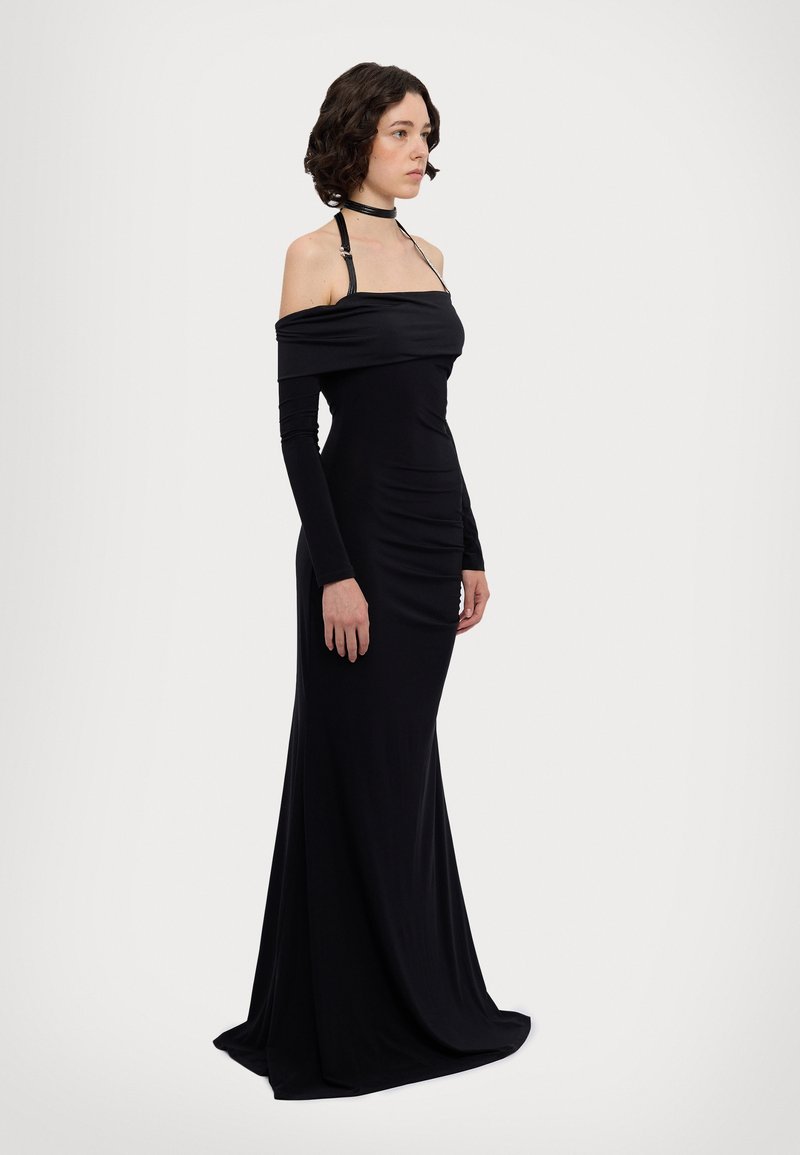 Robe noire décolletée en épaules, à manches longues, avec un tissu drapé et une fente sur le côté. Présente une silhouette ajustée et un design épuré.