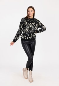 Schwarzer und crèmefarbener gemusterter Pullover mit langen Ärmeln, geripptem Saum, kombiniert mit schwarzen, glänzenden Kunstleder-Hosen und beige Ankle Boots.