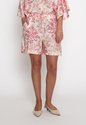 Kvinde iført creme- og lyserøde shorts med mønster og matchende top, beige spidse slingback-højhælede sko og et sølvarmbånd på hendes højre håndled.