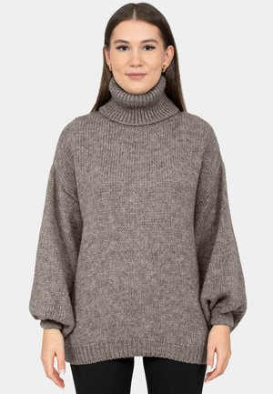 Mujer con un suéter gris holgado de cuello alto y mangas abullonadas, de pie frente a un fondo claro y liso.