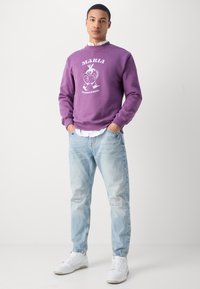 Lilla hettegenser med grafikk på forsiden, rund halslinning og ribbestrikkede mansjetter, sammen med lyseblå denimjeans og hvite sneakers.