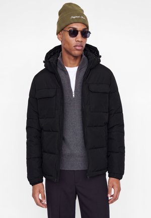 Homme portant une doudoune noire par-dessus un pull gris à zip, un bonnet vert olive avec texte blanc, des lunettes de soleil noires et un pantalon sombre sur fond blanc.