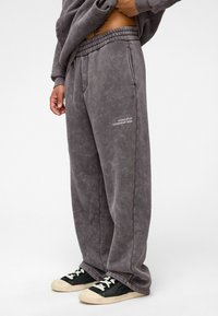 Grijze, oversized sweatpants met een zachte textuur. Bevat een elastische tailleband en witte tekst aan de linkerkant. Draagt met zwarte schoenen.