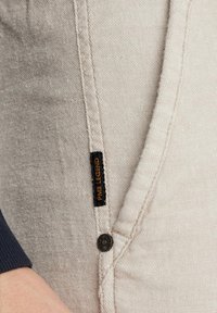 Lichtgrijze broek van textuurstof, met een zwart label met "PANE LEGEND" en een donkere knoopdetail op de zijnaad.