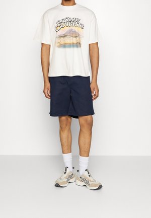 T-shirt grafica color crema con stampa "Glow Lights", abbinata a pantaloni corti blu navy. Calzini alla caviglia bianchi e sneakers beige con accenti neri.