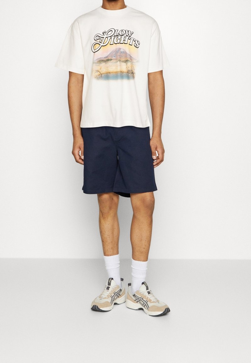 Camiseta gráfica crema con estampado de "Glow Lights", combinada con pantalones cortos azul marino. Calcetines blancos hasta el tobillo y zapatillas beige con detalles en negro.