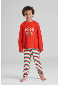 Camisa roja de manga larga con el texto "MINI ELF" en blanco y verde, combinada con pantalones blancos que tienen rayas rojas y verdes.