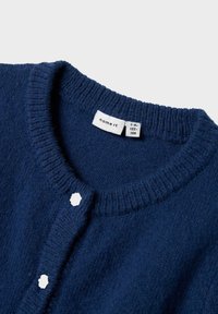 Maglione lavorato a maglia di colore blu scuro con colletto rotondo e bottoni bianchi a forma di fiore, etichetta della taglia che indica 7-8 anni, 122-128 cm.