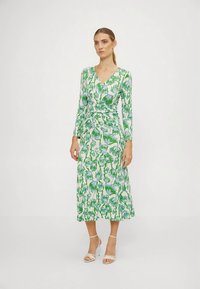 Kobieta stoi w zielono-białej kwiatowej sukience midi z długim rękawem i dekoltem w kształcie litery V, w białych sandałach na obcasie, na jednolitym tle.