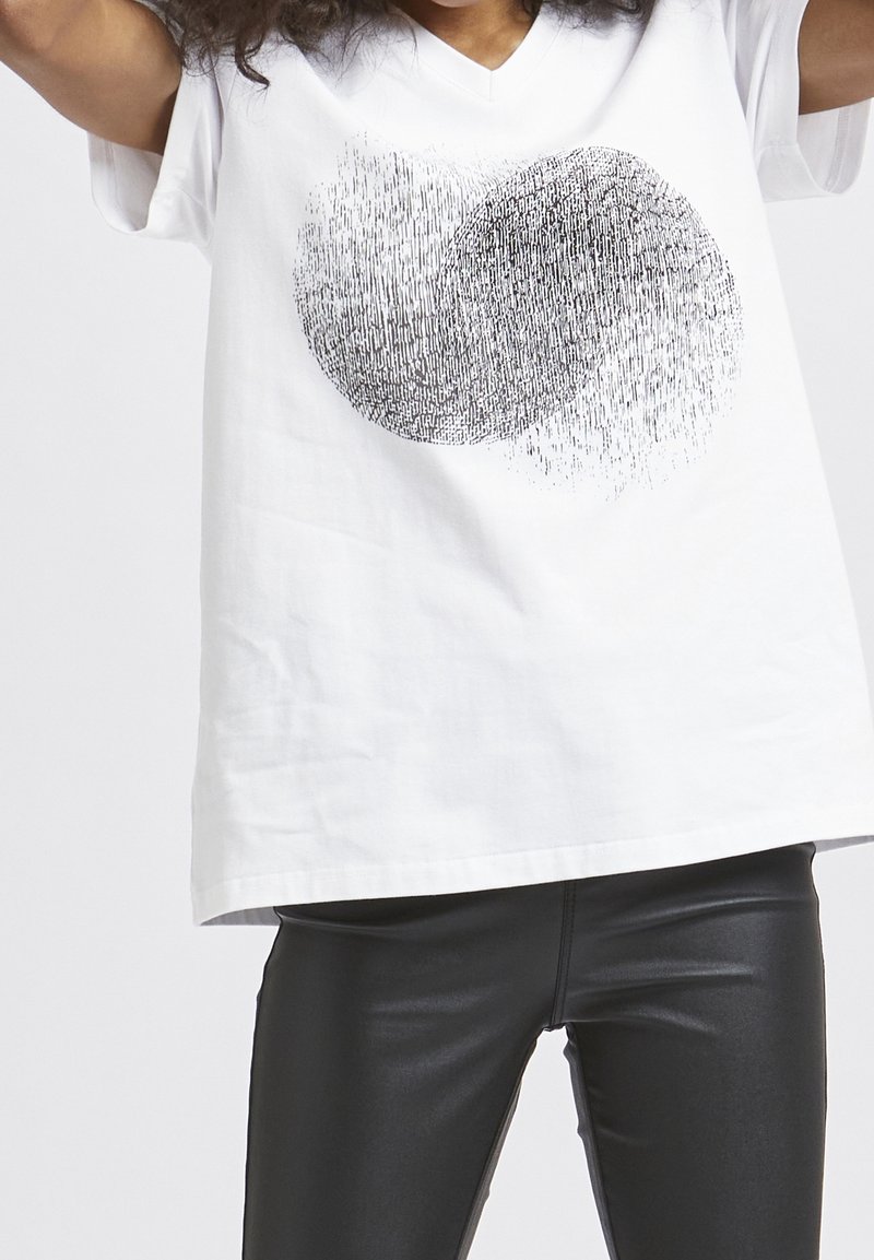 Personne portant un t-shirt blanc à col en V avec un motif circulaire abstrait noir et un pantalon en cuir noir sur un fond uni.