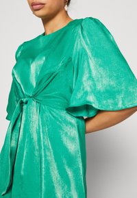 Femme portant une robe verte émeraude brillante avec des manches courtes bouffantes et un détail de nœud à l'avant, assortie d'une unique boucle d'oreille pendante en perle.