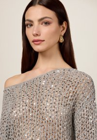 Zilveren haaktop met een off-shoulder ontwerp, voorzien van sprankelende pailletten, een losse draping en een gestructureerd breipatroon.