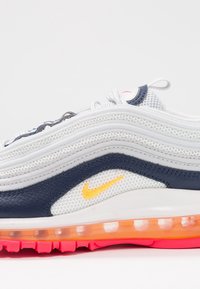 Der Sneaker verfügt über ein Obermaterial aus weißem Mesh mit navyblauen Lederelementen, einer orangefarbenen Air-Unit-Innensohle, einer pinken Gummisohle und einem gelben Logodetail.