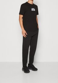 Camiseta de algodón negra con un parche de logo Calvin Klein blanco, combinada con pantalones negros y zapatillas deportivas negras. Diseño simple y casual.