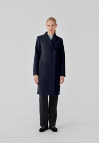 Námořnický kabát z vlněné směsi s dvojitým zapínáním, strukturovanými klopami, bočními kapsami a designem ke kolenům, stylizovaný přes tmavé kalhoty.