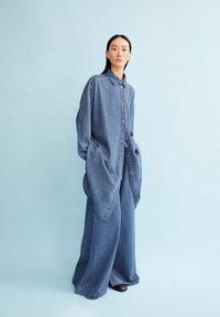 Hellblaue Denim-Bluse in Übergröße mit durchgehender Knopfleiste, langen Ärmeln und abgerundeten Säumen. Kombiniert mit weit geschnittenen Denim-Hosen.