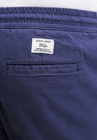 Achteraanzicht van marineblauwe broek met elastische tailleband, een enkele achterzak en een wit label met de tekst "Pier One Comfort & Quality Premium."