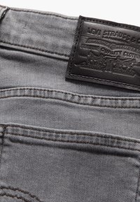 Siva denimska tkanina z teksturirano površino, ki vključuje črn usnjen obliž z embossiranim logotipom blagovne znamke in kontrastnim šivanjem.