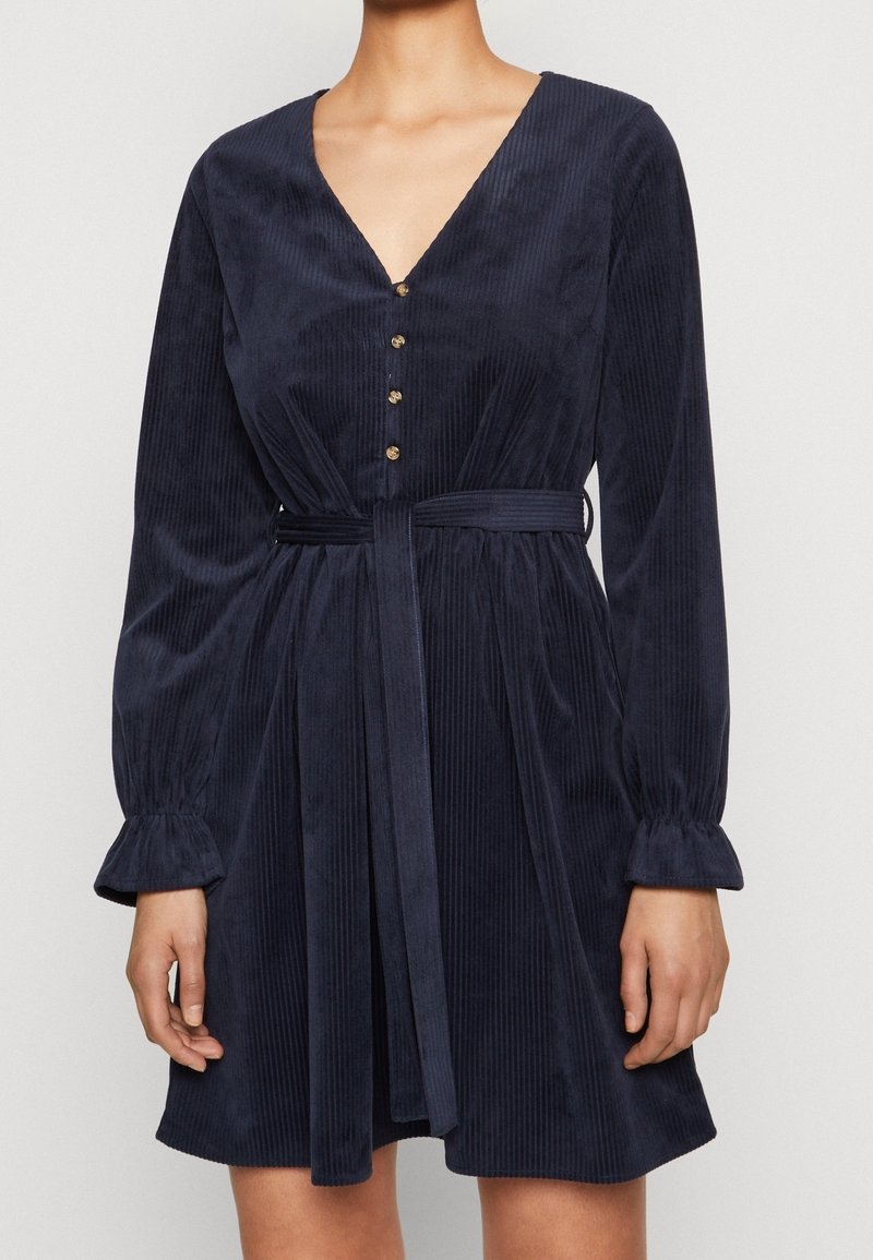 Femme portant une robe en velours côtelé bleu marine à manches longues, boutons dorés sur le devant, décolleté en V et ceinture en tissu assorti.
