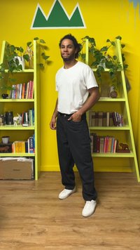 Uma pessoa vestindo uma t-shirt branca e calças cargo pretas está encostada numa parede amarela com formas de montanhas verdes e prateleiras verdes com plantas.