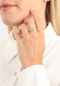 Anello intrecciato d'oro con accenti verdi e gialli indossato su una mano appoggiata vicino al viso. La persona indossa una camicia bianca con colletto.