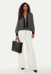 Donna che indossa una blusa nera a fantasia, pantaloni bianchi a gamba larga, scarpe nere a punta, mentre tiene in mano una borsa Guess nera, in piedi davanti a uno sfondo semplice.