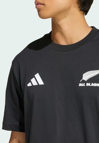 Czarna bawełniana koszulka z okrągłym dekoltem, z białym logo Adidas oraz napisem "ALL BLACKS" i grafiką paproci na klatce piersiowej.