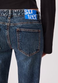Jean en denim bleu avec une texture légèrement décolorée, présentant des coutures orange, deux poches arrière et une étiquette bleue avec "SLI teen" au niveau de la taille.