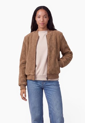 Donna che indossa una giacca bomber in pile marrone, maglione beige e jeans blu su sfondo bianco uniforme.