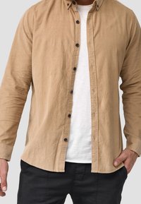 Langärmeliges, beige Hemd mit Button-Down-Kragen, durchgehender Knopfleiste und lockerem Schnitt, kombiniert mit einem weißen T-Shirt darunter.