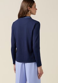 Mujer de perfil vistiendo una camiseta de manga larga azul marino, pantalones anchos de pierna en azul claro y pendientes geométricos dorados sobre un fondo beige.