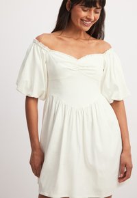 Vit off-shoulder klänning med puffärmar, med en flätad byst och en utsvängd kjol. Tillverkad av lätt, strukturerad tyg.