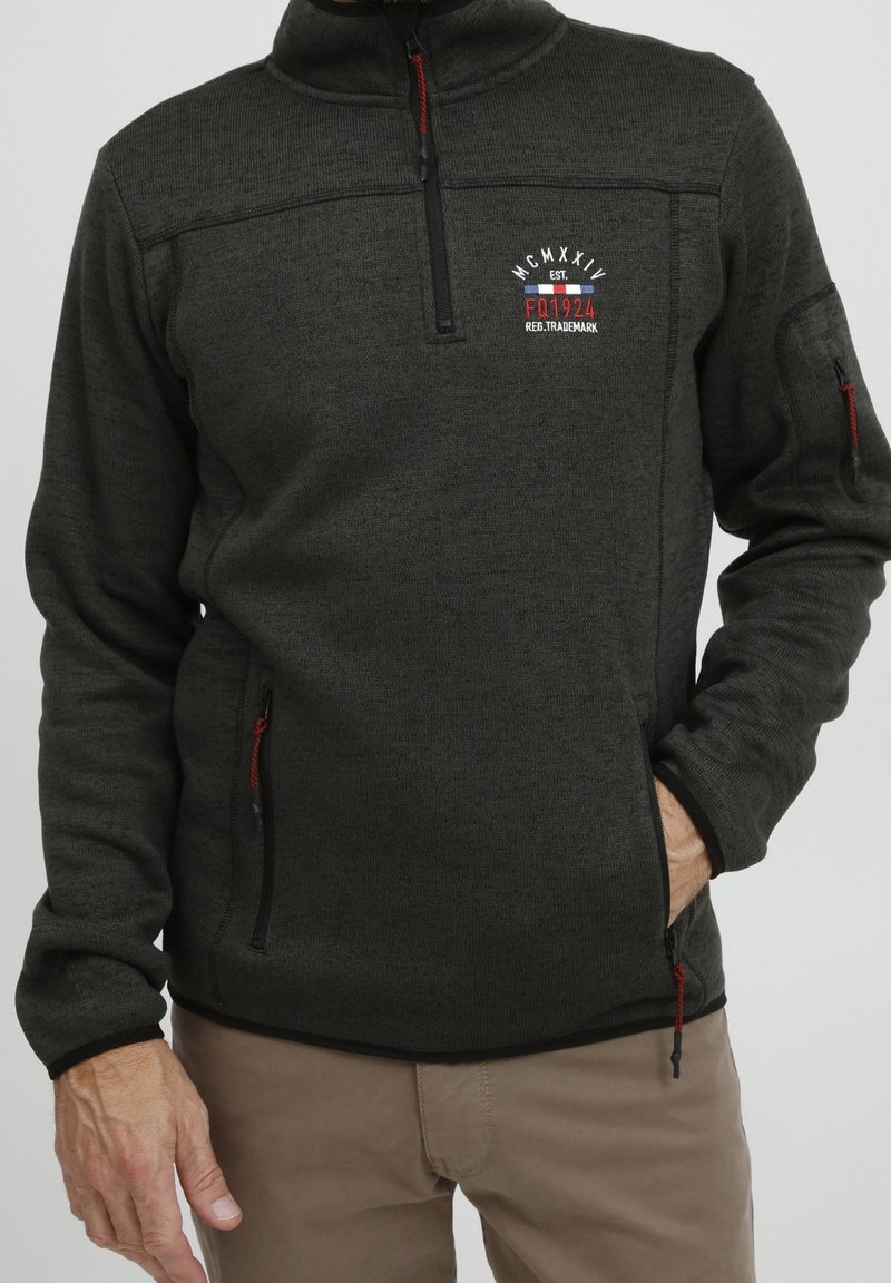 Polaris gris foncé à zip quart avec curseurs rouges, logo sur la poitrine gauche, poches zippées, porté avec un pantalon beige, une main dans la poche.