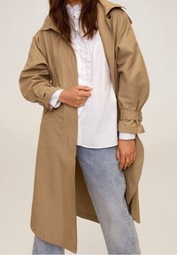 Manteau beige en trench avec une coupe décontractée, des poignets boutonnés et une capuche. Porté par-dessus un chemisier blanc à volants et un jean bleu clair.